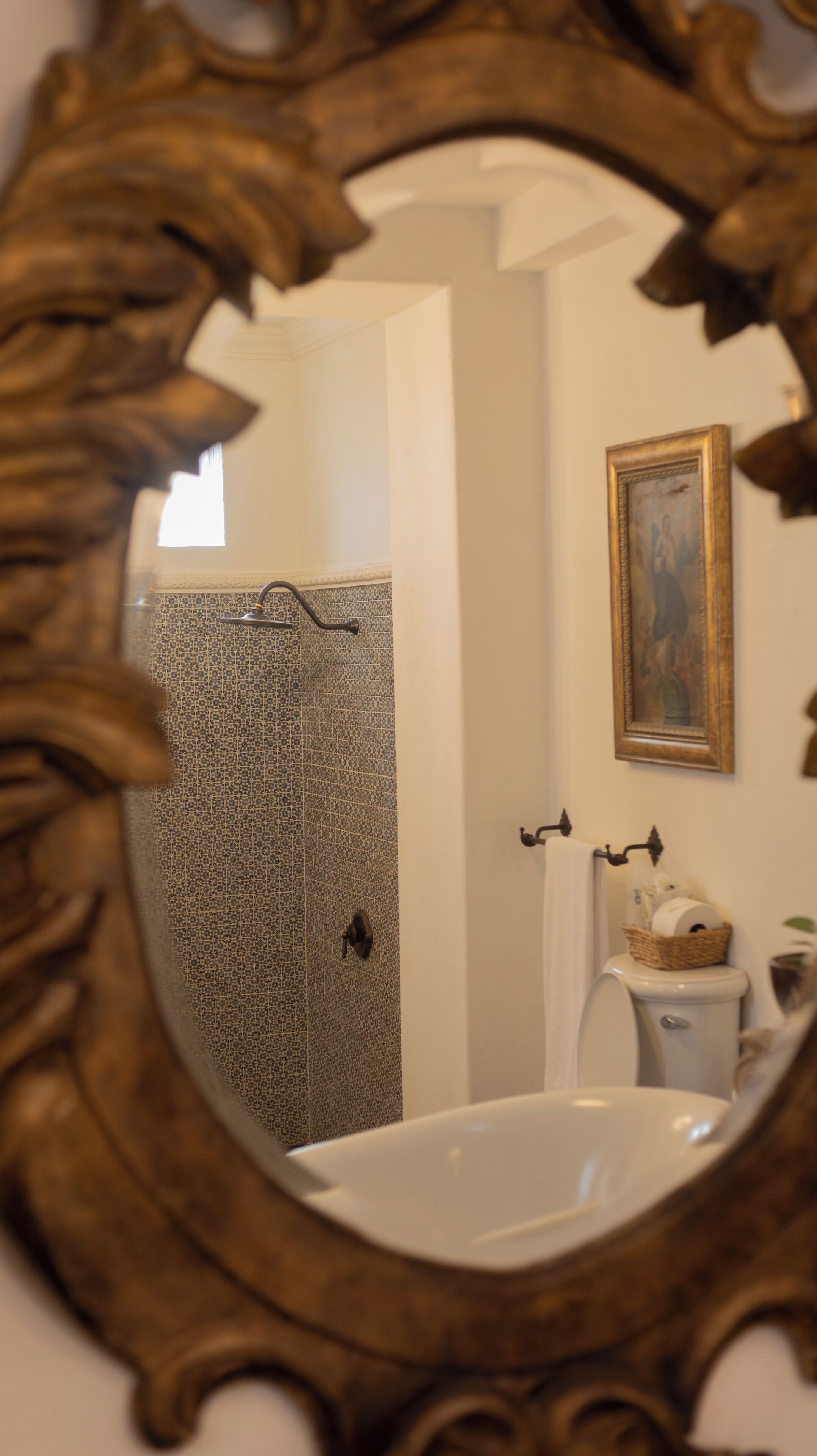 A bathroom with a white sink, a shower with patterned tiles, a framed painting on the wall, and a toilet with a basket of toiletries.