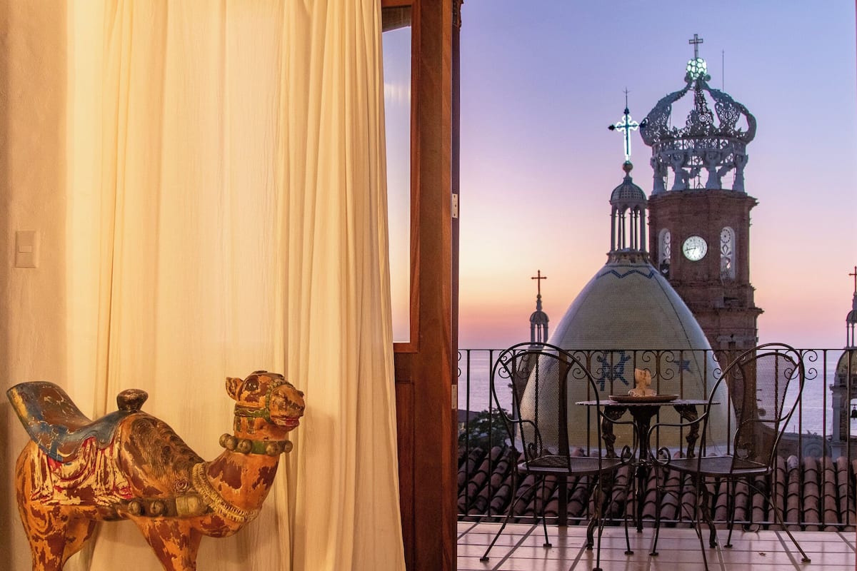 A balcony with a view of a historic church with a clock tower and crosses, a camel-shaped chair, and a table with chairs.