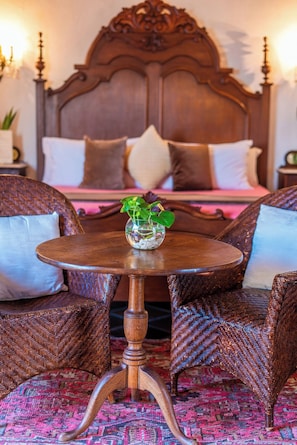 A bed with a wooden headboard, two wicker chairs, a round wooden table with a glass vase, and a patterned carpet.