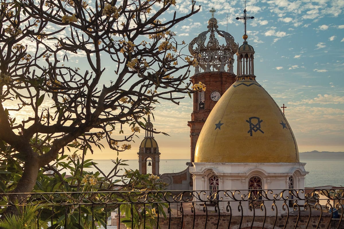 A church with a golden dome and a bell tower, surrounded by trees and overlooking the sea.
