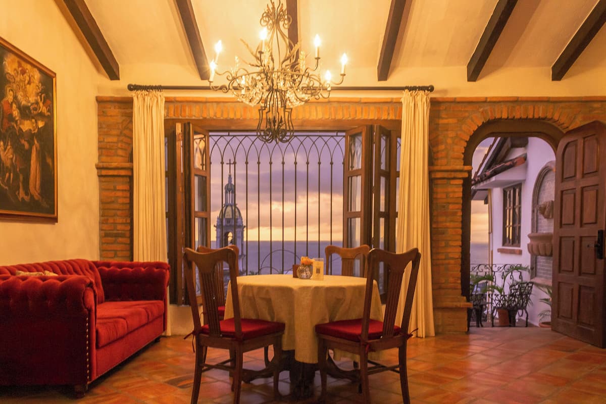 A room with a red sofa, a table with chairs, a chandelier, and a view through a window with a metal gate.