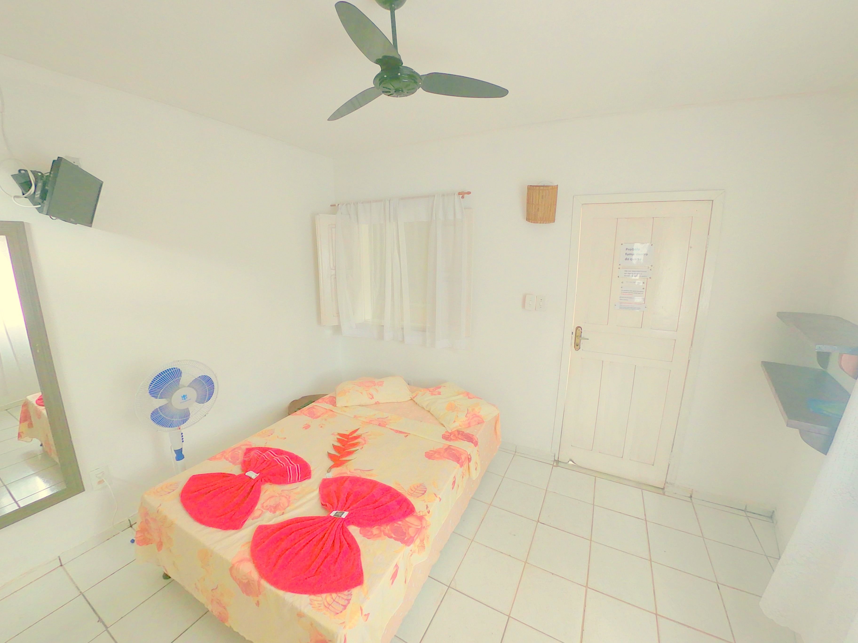 A room with a bed, a ceiling fan, a window with curtains, a door, and a mirror.