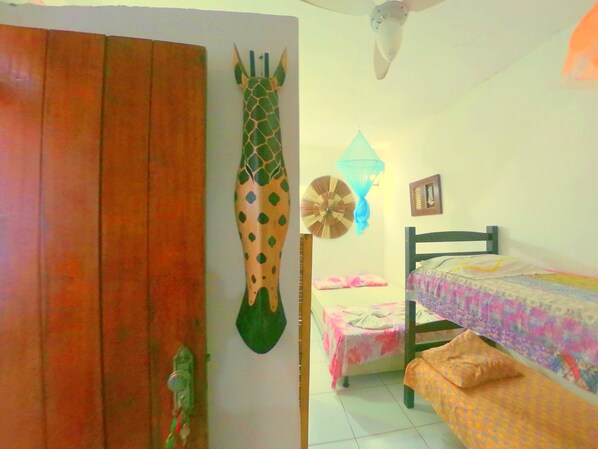 A room with a wooden door, a giraffe decoration, a bed with a colorful bedspread, a small table with a pink tablecloth, and a wall-mounted fan. - Casa da Pituba (Itacaré)