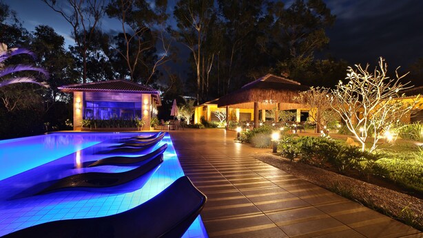 Una piscina con iluminación azul, una terraza de madera y un edificio con techo de paja.