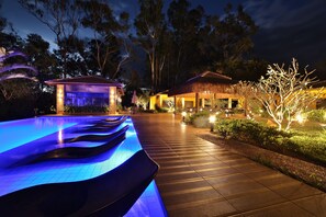 Una piscina con iluminación azul, una terraza de madera y un edificio con techo de paja.