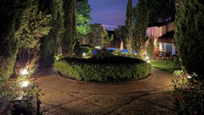 Un jardín nocturno con un camino pavimentado, altos árboles y una zona de piscina.