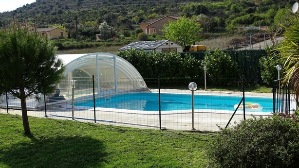 A swimming pool with a glass enclosure, surrounded by a fence, and set against a hillside with houses in the background. - Soleil single-storey villa with 8/4 saltwater swimming pool, secure, fenced garden  (Lagorce)