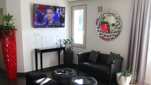 A modern living room with a black leather sofa, a coffee table, and a TV mounted on the wall. There are potted plants and a decorative mirror on the wall. - Doubs Hotel - Hostel (Ecole-Valentin)