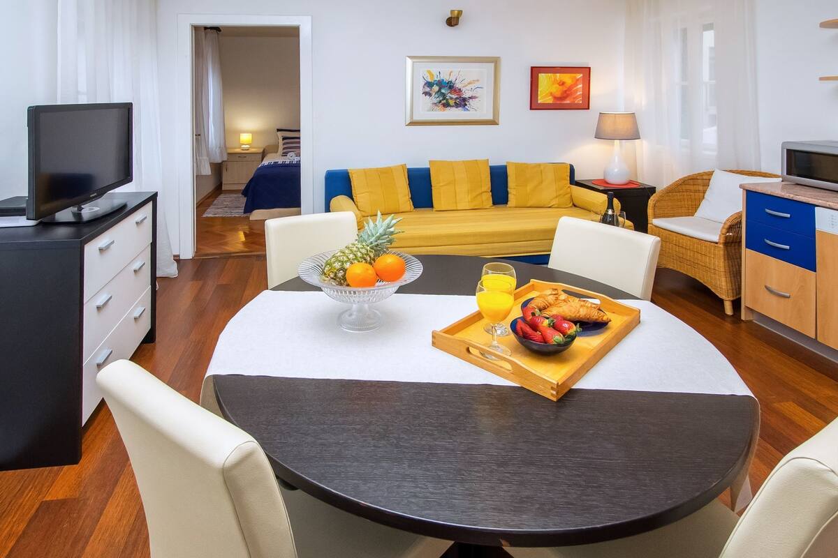 A modern living room with a round dining table, a sofa with yellow cushions, a television, and a dining area with breakfast items.