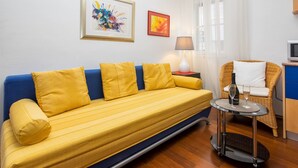 A yellow sofa with blue cushions, a wicker chair, a small table with a bottle and glass, and two framed artworks on the wall. - Guest House Leta (Split)