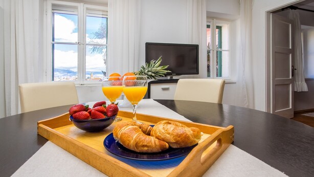 A breakfast tray with croissants, strawberries, and orange juice on a table in a hotel room with a TV and a view of the city. - Guest House Leta (Split)
