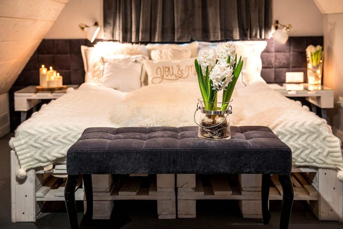 A cozy bedroom with a bed, a bedside table with candles, a nightstand, and a decorative vase with flowers.