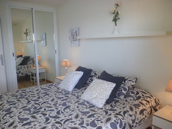 A bedroom with a bed, two bedside tables, a nightstand, a mirror, and a wall-mounted shelf with a vase.