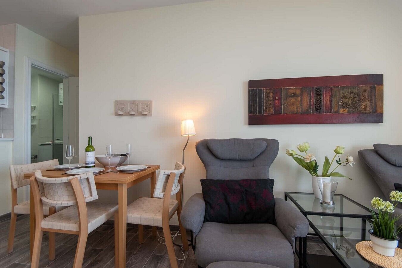 A modern living room with a dining area, a grey armchair, a glass coffee table, and a painting on the wall.