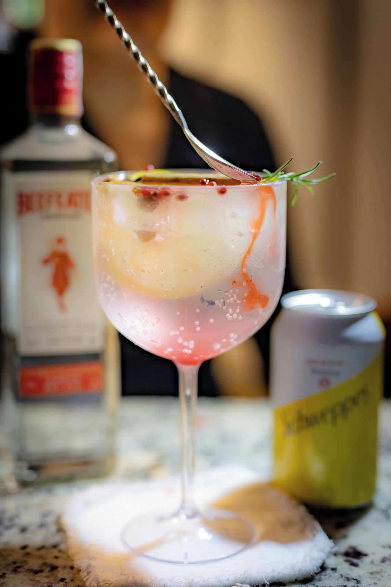 A cocktail glass with a straw, garnished with a sprig of rosemary, on a marble surface. Behind it, there is a bottle of liquor and a can of soda.