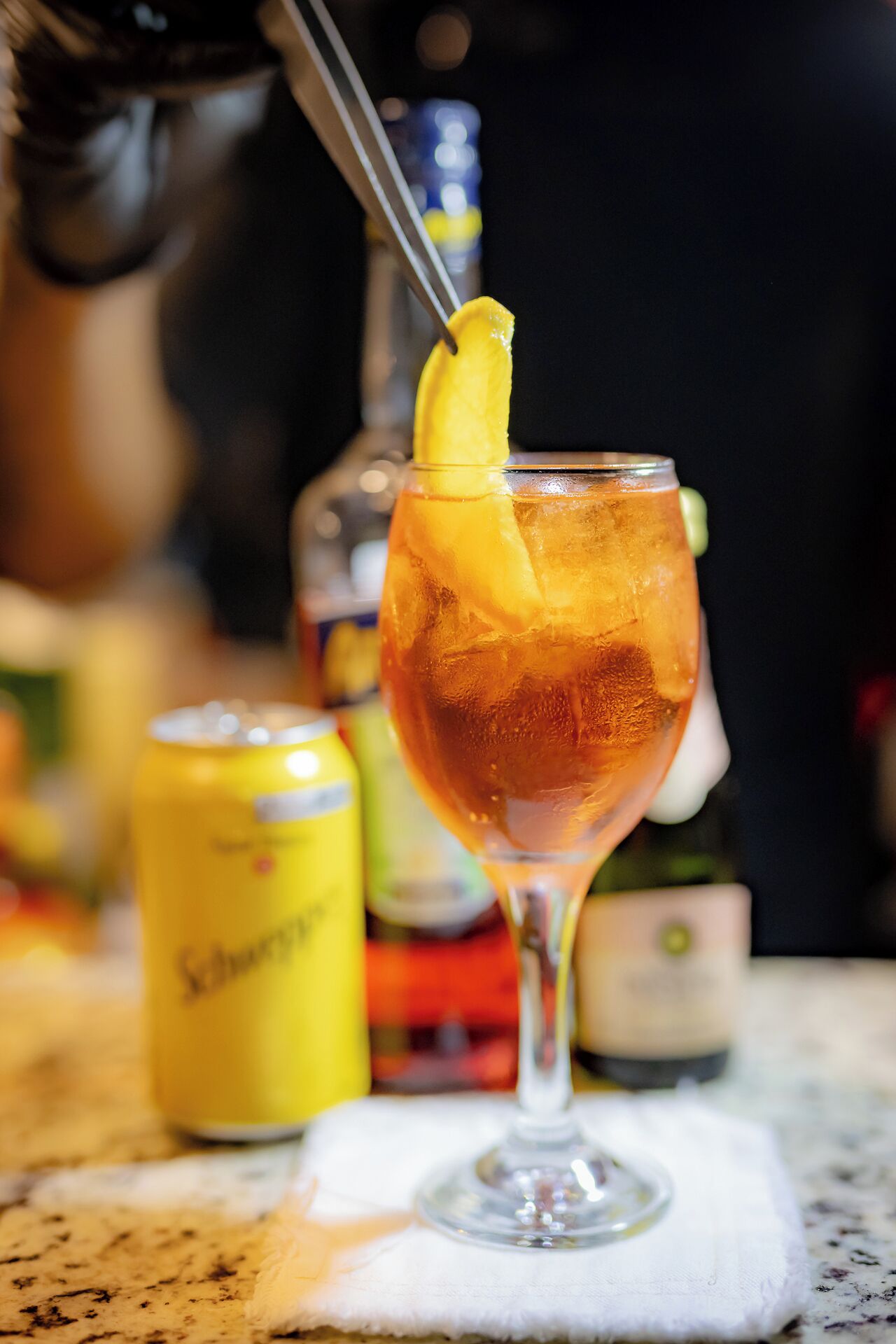 Un cocktail guarnito con una fetta di limone, una lattina di Schweppes e una bottiglia sullo sfondo.