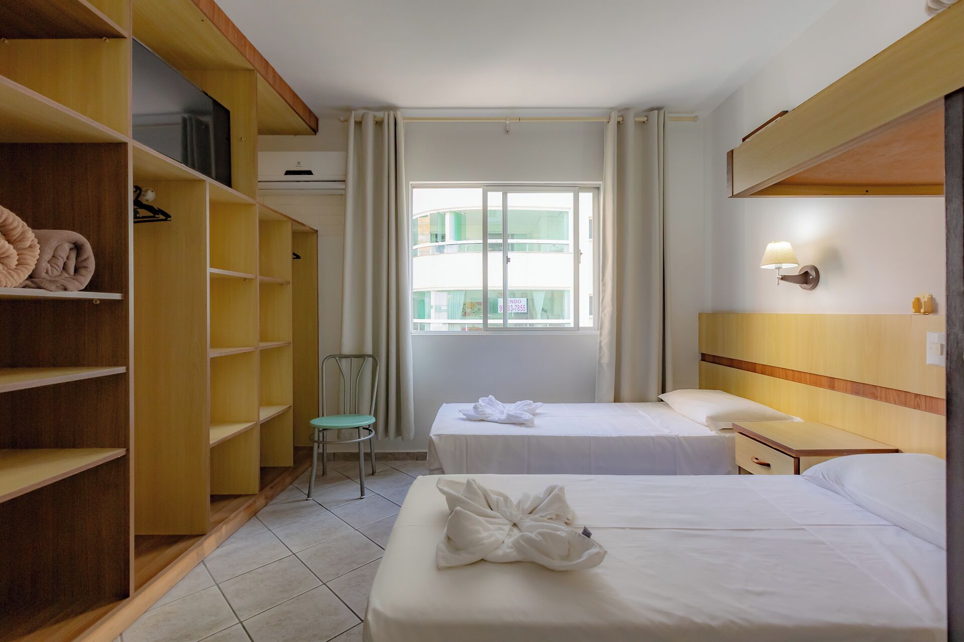 Habitación de hotel con dos camas, una silla, un armario y una ventana con vistas a edificios.