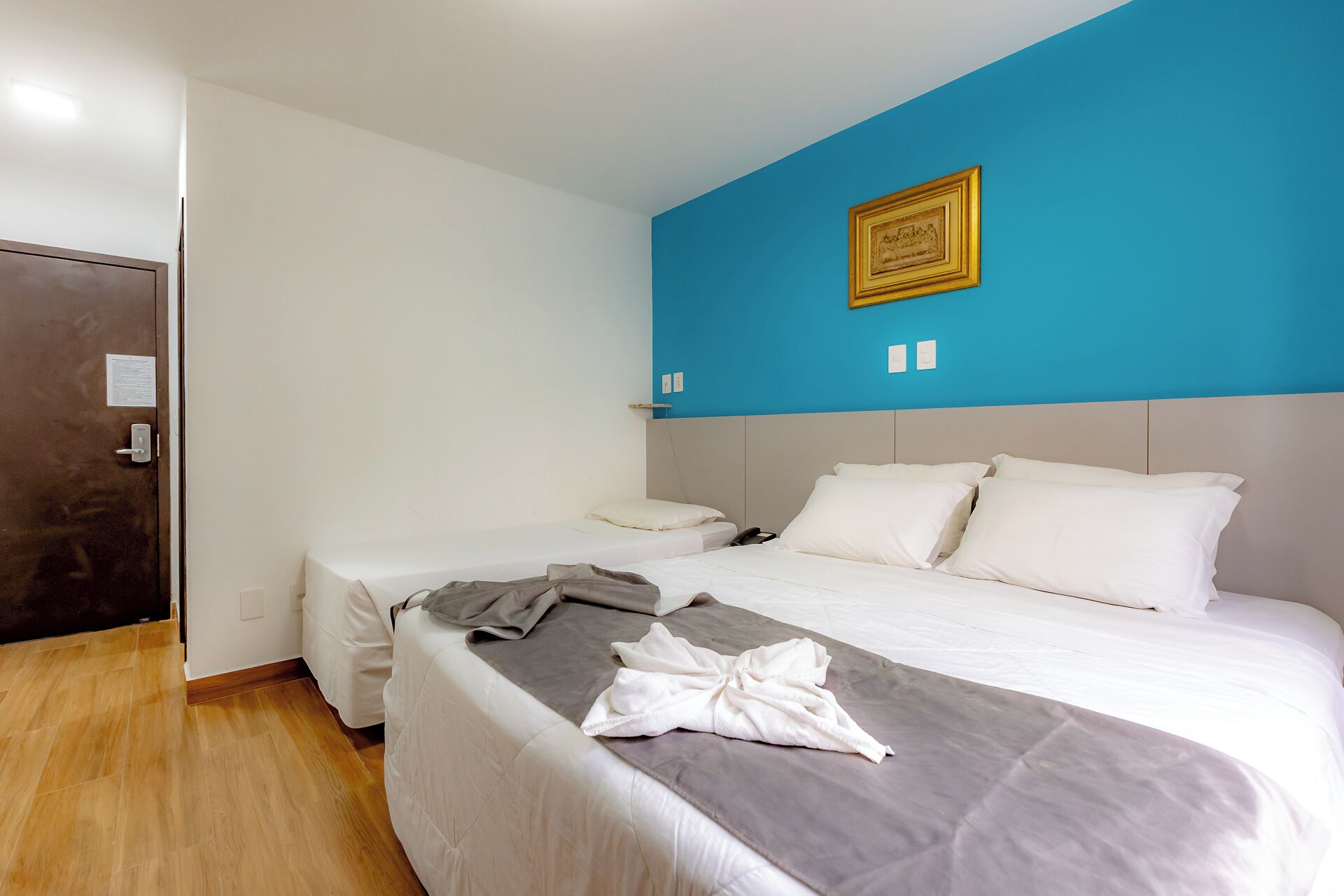 A hotel room with a large bed, two smaller beds, a wooden floor, a blue accent wall, and a framed picture.