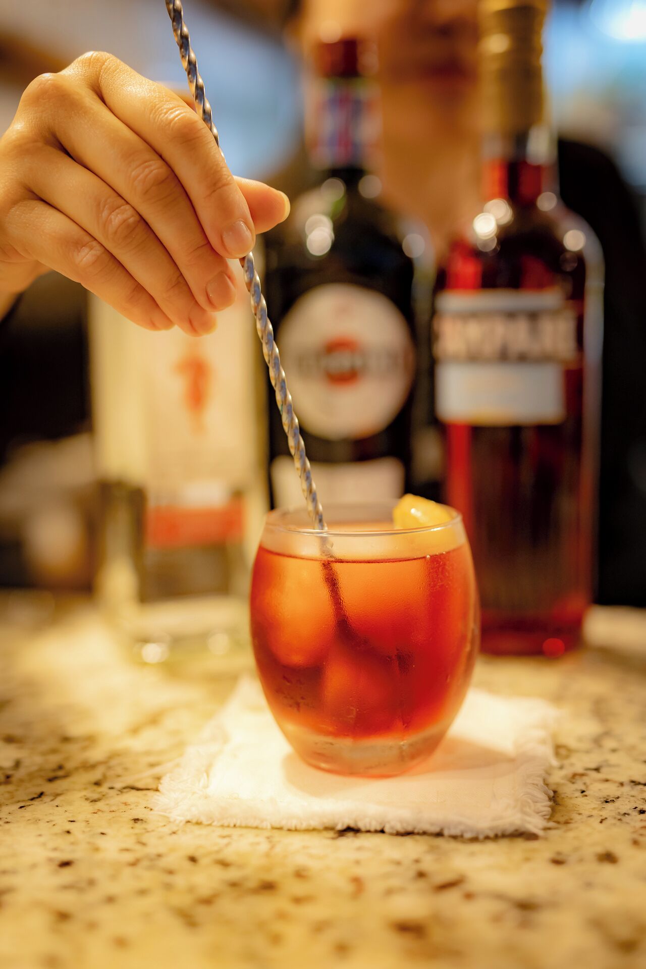 A hand stirring a cocktail in a glass with ice cubes, with two bottles of liquor in the background.