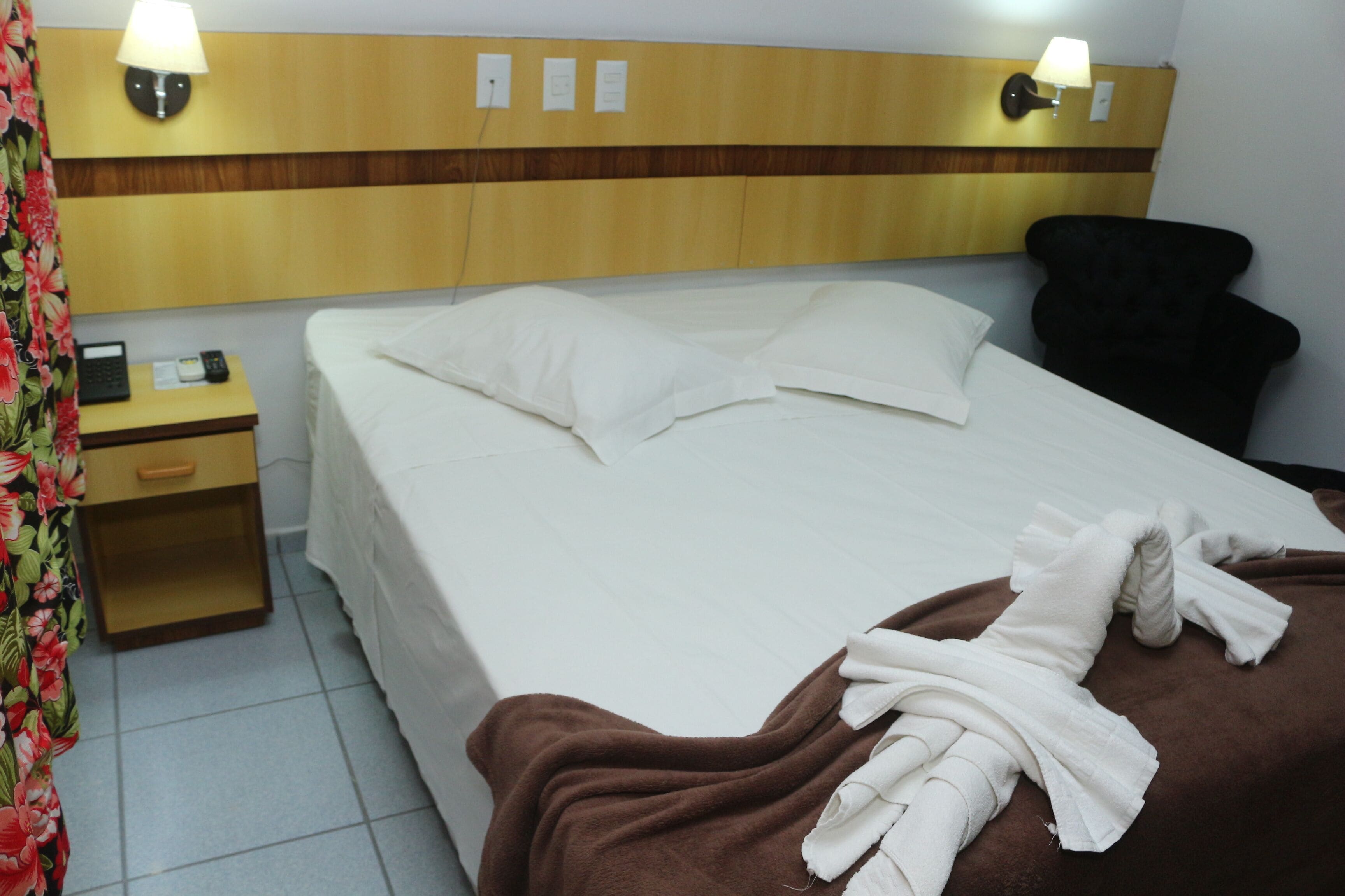 A hotel room with a bed, a nightstand with a phone and a lamp, and a brown blanket on the bed.