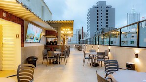 Un espacio de comedor al aire libre con mesas y sillas, una pequeña barra y vistas al horizonte de la ciudad.