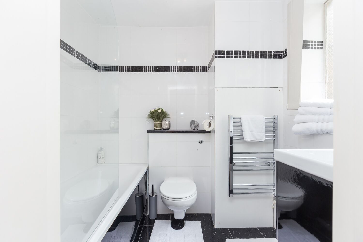 Een moderne badkamer met een wit toilet, een glazen douchecabine, een zwart-wit betegelde vloer en een wit wastafelmeubel met een handdoekhouder.