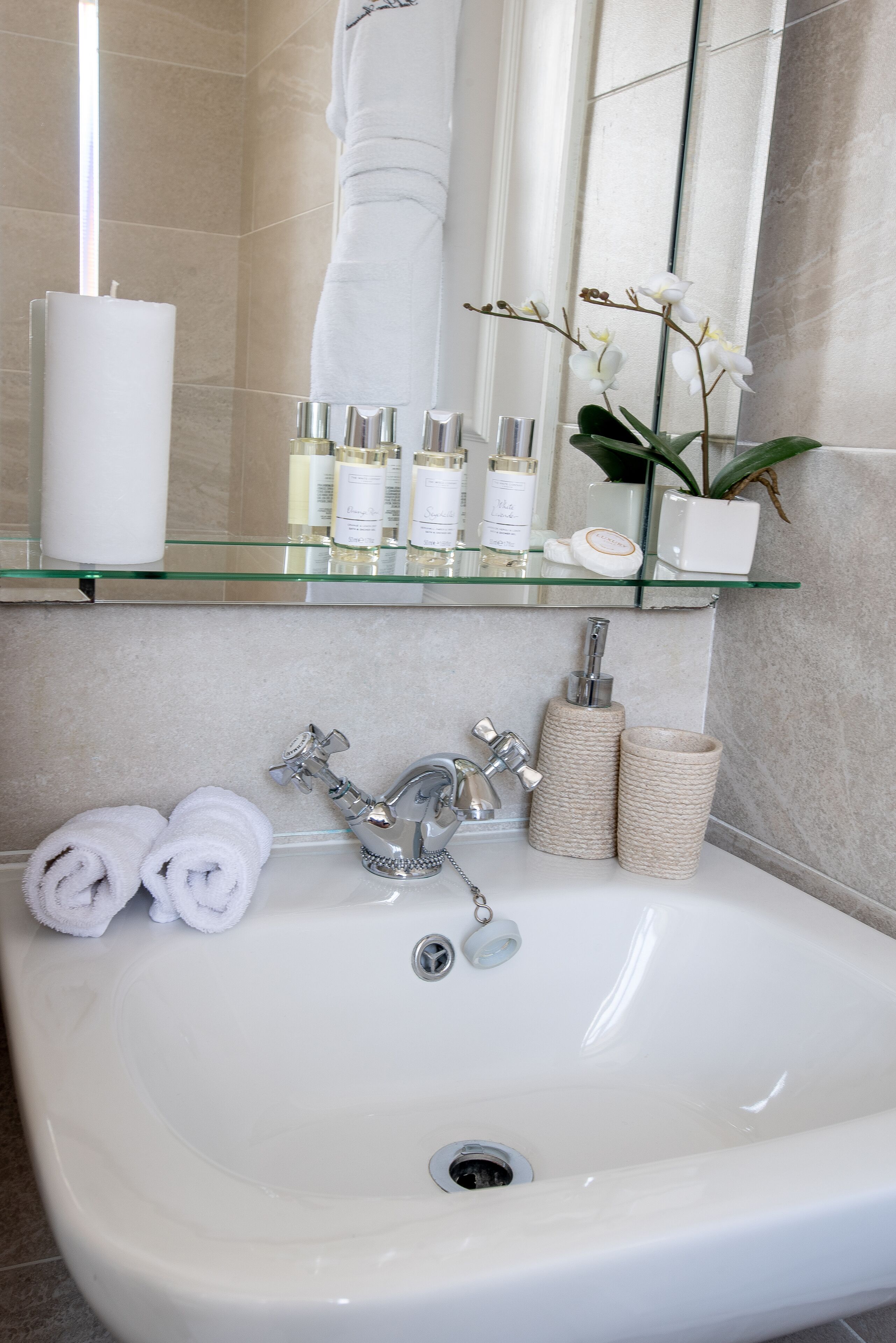 Een badkamer met een witte wasbak, twee opgerolde handdoeken, een glazen plank met toiletartikelen en een potorkidee.