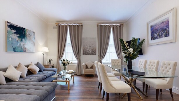 Een moderne woonkamer met een grijze bank, een glazen salontafel, en een eethoek met een glazen tafel en witte stoelen.