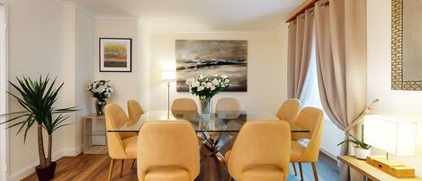 A dining area with a glass table, four chairs, a vase of flowers, and a painting on the wall.