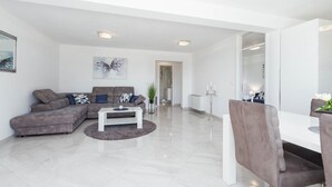 Un salon moderne avec un canapé d’angle gris, une table basse blanche et un coin repas comprenant une table et des chaises blanches.