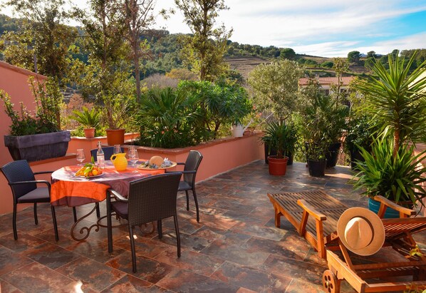 An outdoor patio with a table set for dining, surrounded by potted plants and a view of the countryside.