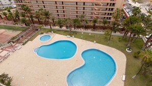 Un'ampia piscina all'aperto con diverse sezioni, circondata da palme e un edificio a più piani sullo sfondo.