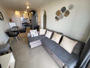 Uma sala de estar moderna com um sofá seccional cinza, uma mesa de jantar com cadeiras e uma cozinha ao fundo.