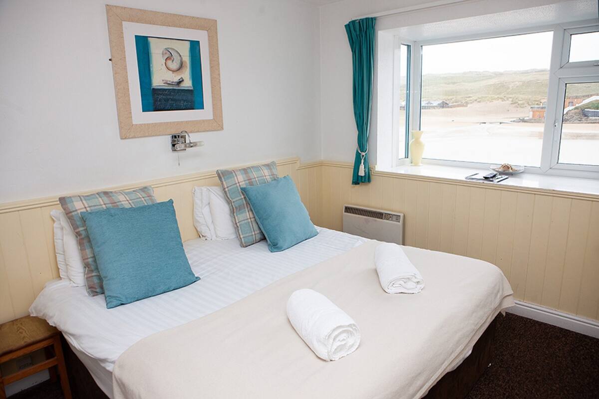 Quarto de hotel com cama, janela com vista, quadro na parede e pequena mesa com abajur.