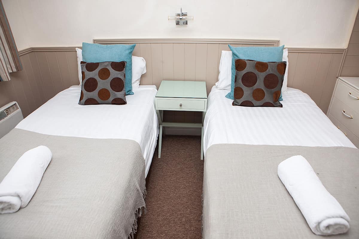 Quarto de hotel com duas camas de solteiro, cada uma com almofada e toalha enrolada. Há uma pequena mesa entre as camas.