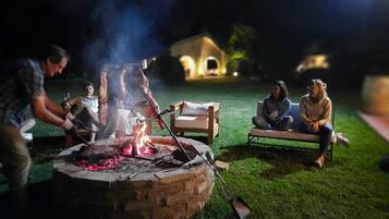 Eine Gruppe von Menschen versammelt sich in der Nacht an einem Lagerfeuer, im Hintergrund sieht man ein Haus.