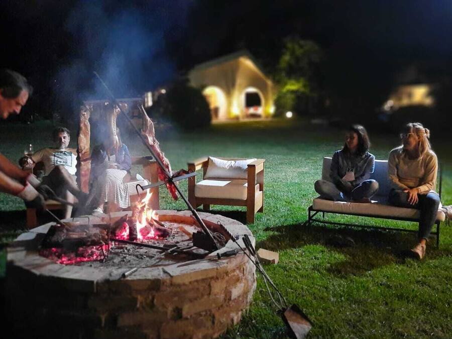 Eine Gruppe von Menschen versammelt sich in der Nacht an einem Lagerfeuer, im Hintergrund sieht man ein Haus.