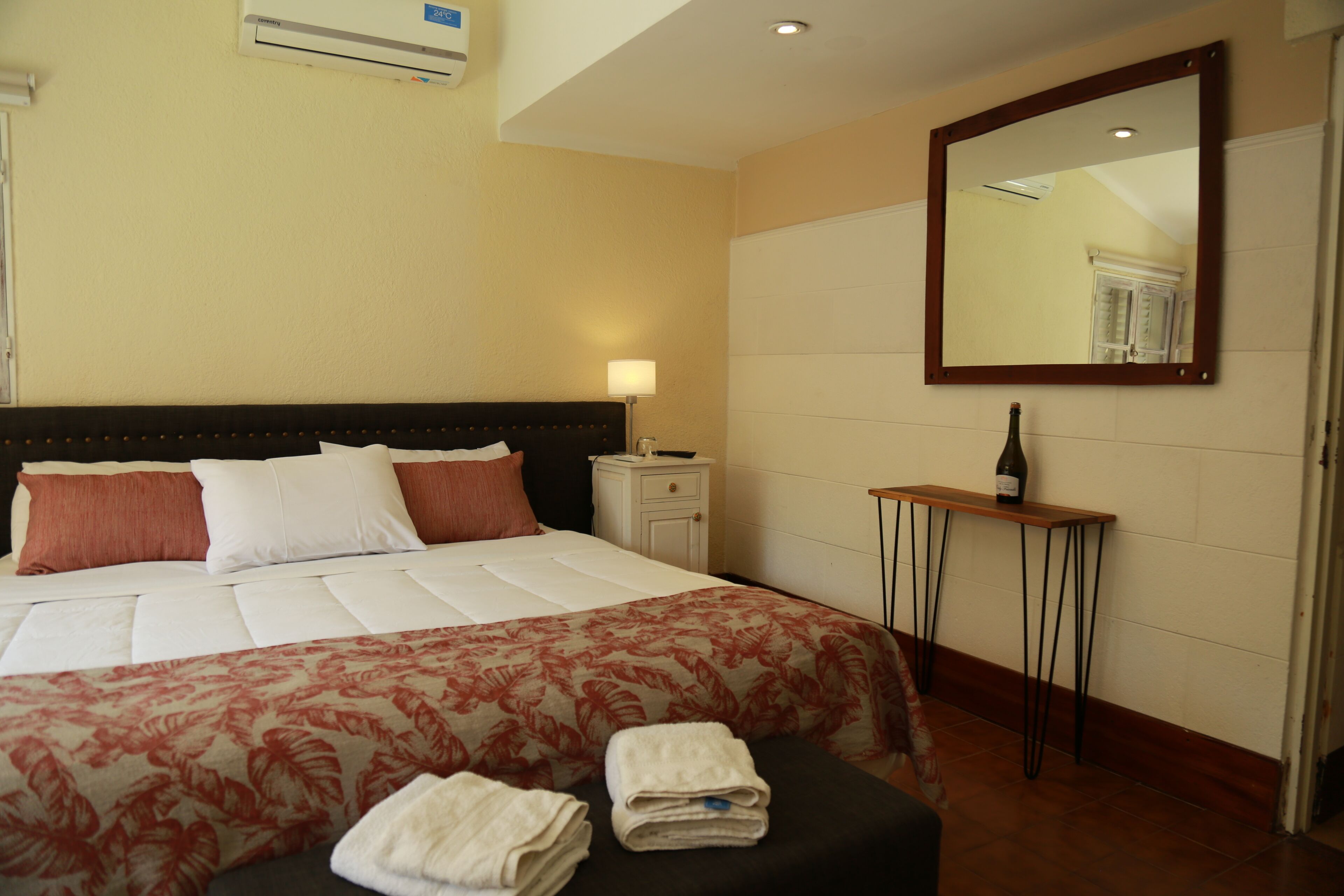 Een hotelkamer met een groot bed, een nachtkastje met een lamp, een kleine tafel met een fles en een spiegel aan de muur.
