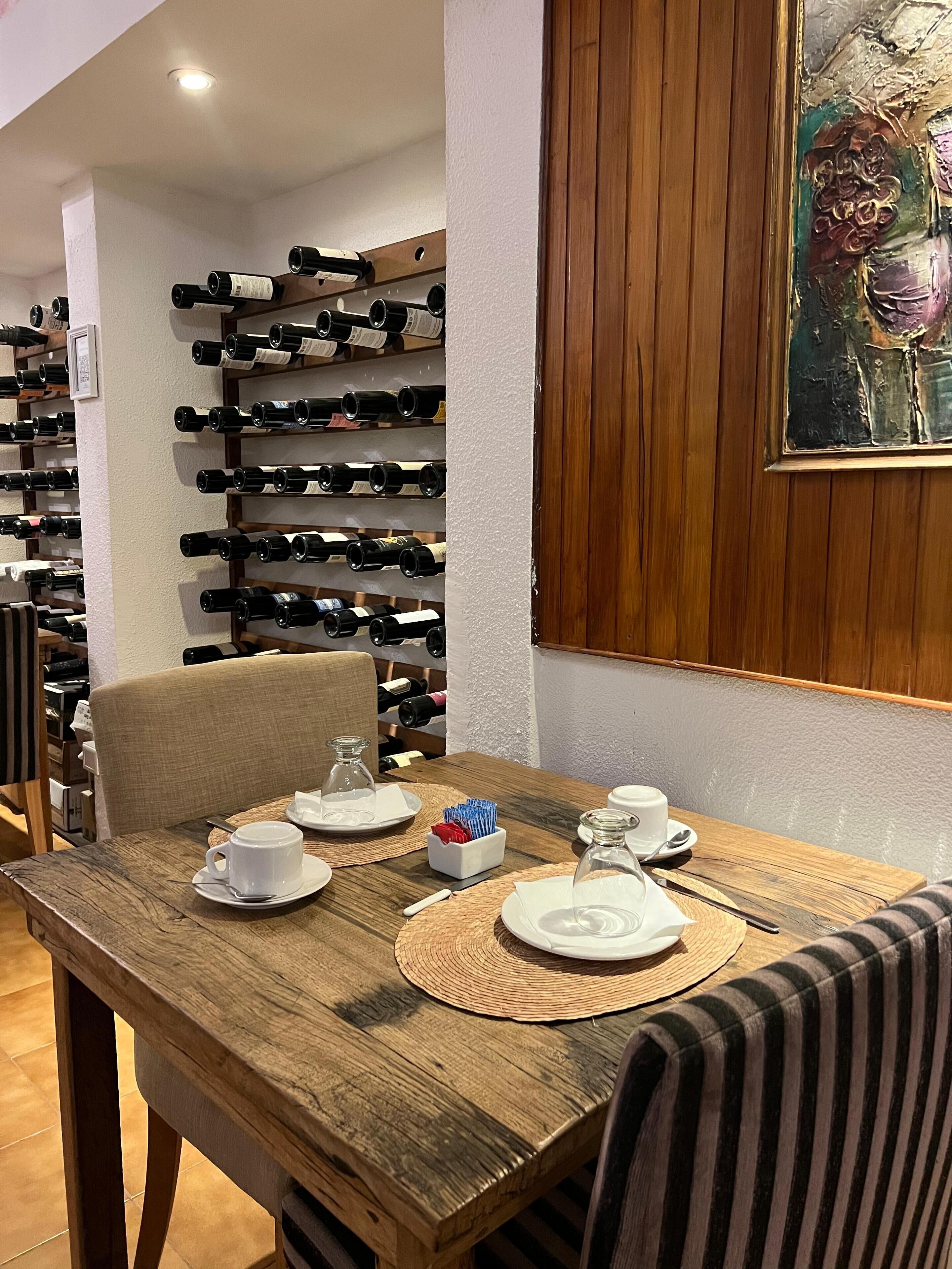 Un coin repas avec une table en bois dressée pour deux personnes, un meuble à vin en arrière-plan et un tableau accroché au mur.