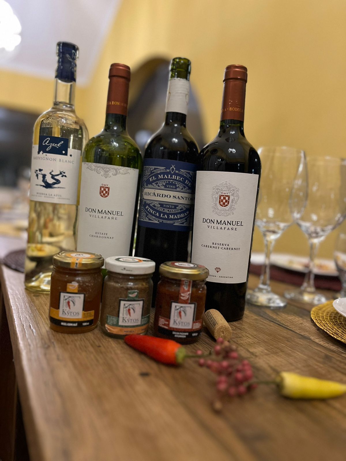 A selection of wine bottles and jars on a wooden table, with glasses and a placemat in the background.