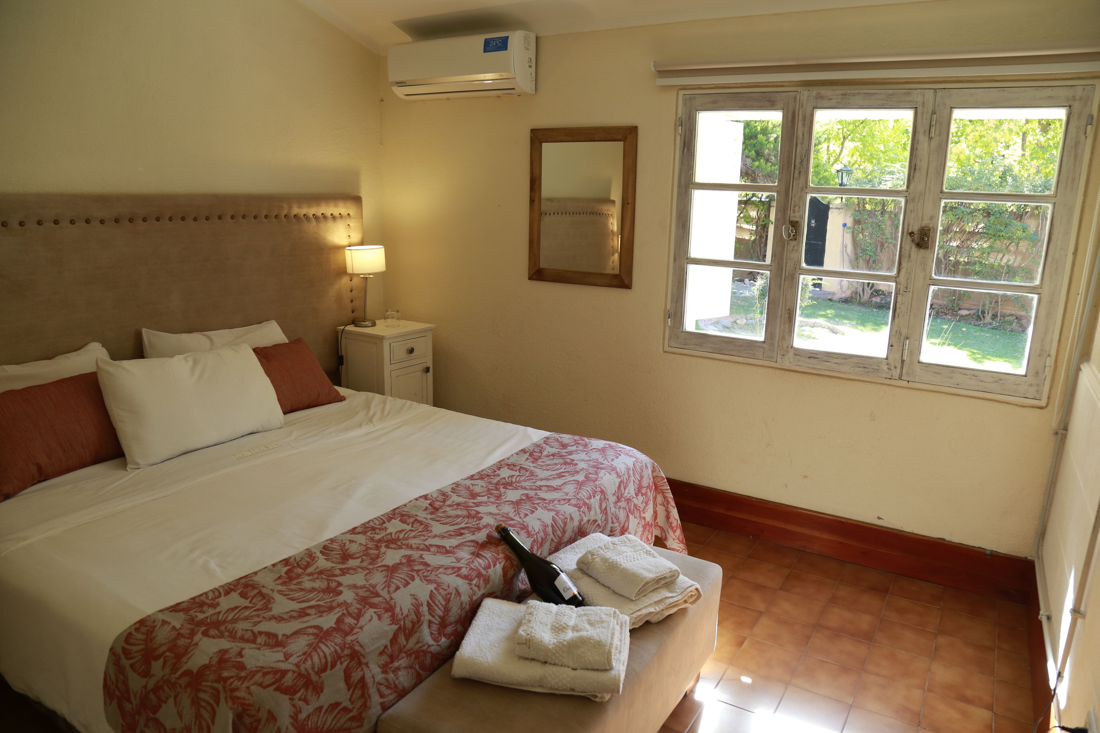 Une chambre d’hôtel avec un grand lit, une table de chevet, un miroir et une fenêtre donnant sur un paysage verdoyant.