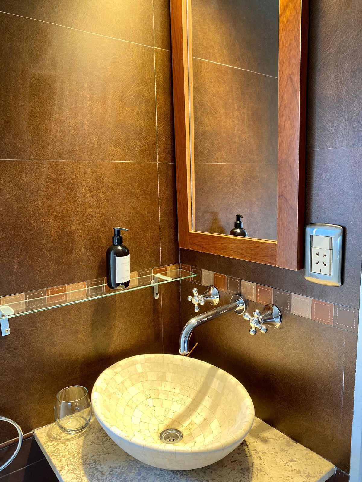 A bathroom with a large mirror, a vessel sink, a glass shelf with a bottle, and a towel rack.
