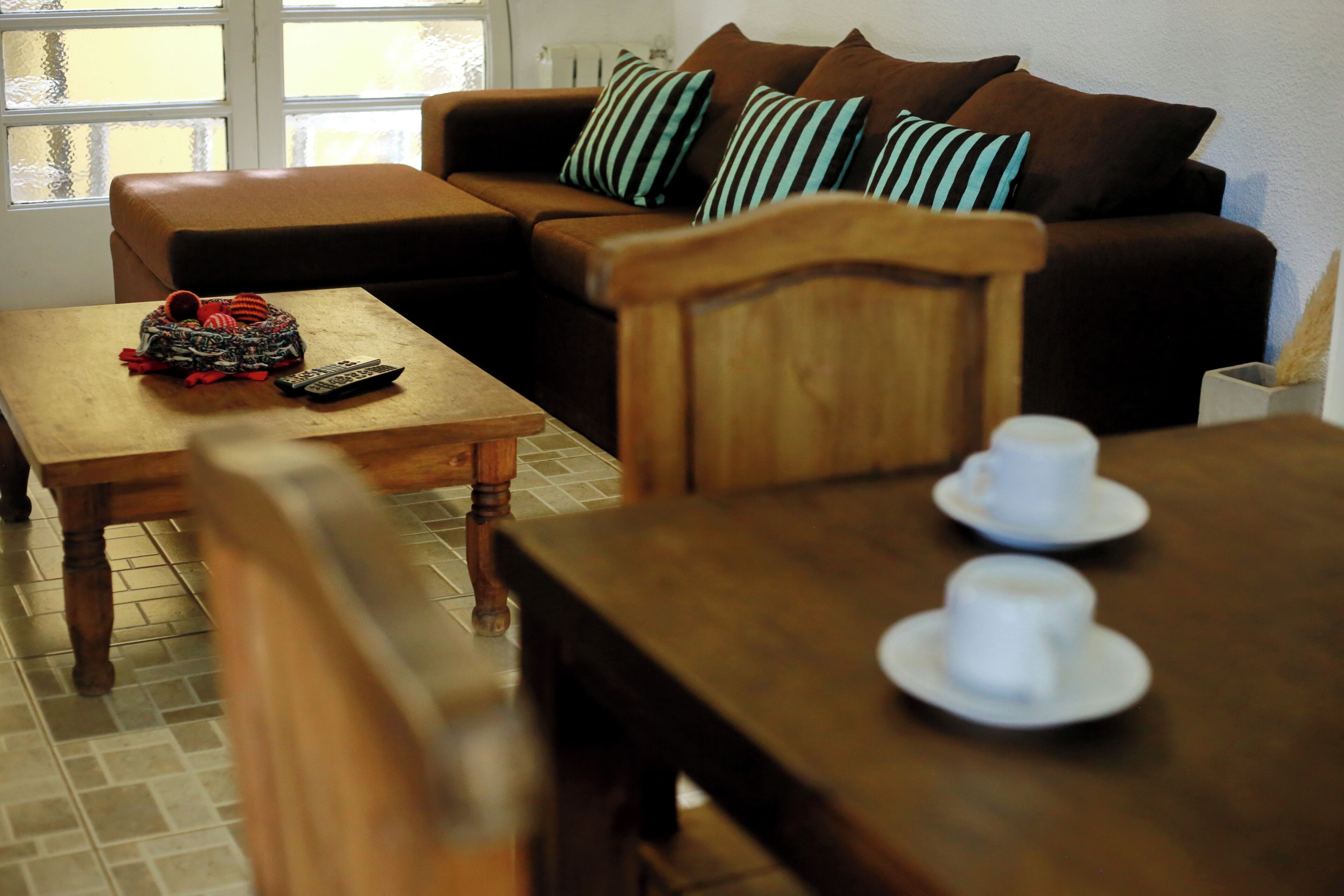 Un salón con un sofá marrón, almohadas a rayas, una mesa de centro de madera y una mesa de comedor con tazas blancas.