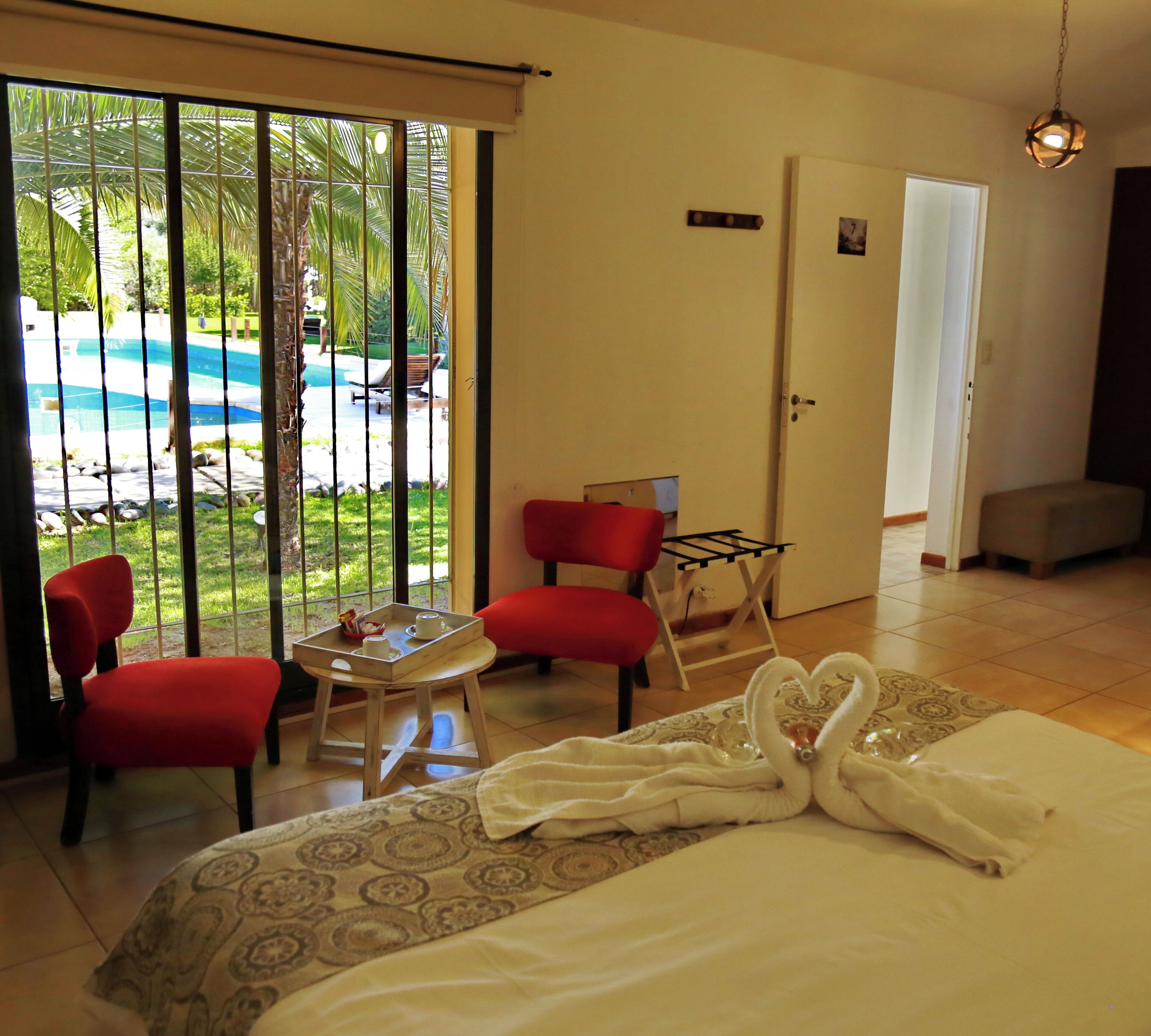 Habitación de hotel con cama, dos sillas rojas, una mesita con bandeja y vista a una piscina a través de un ventanal corredero.