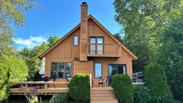 Una casa de madera de dos pisos con balcón, ventanas amplias y una terraza con mobiliario exterior.
