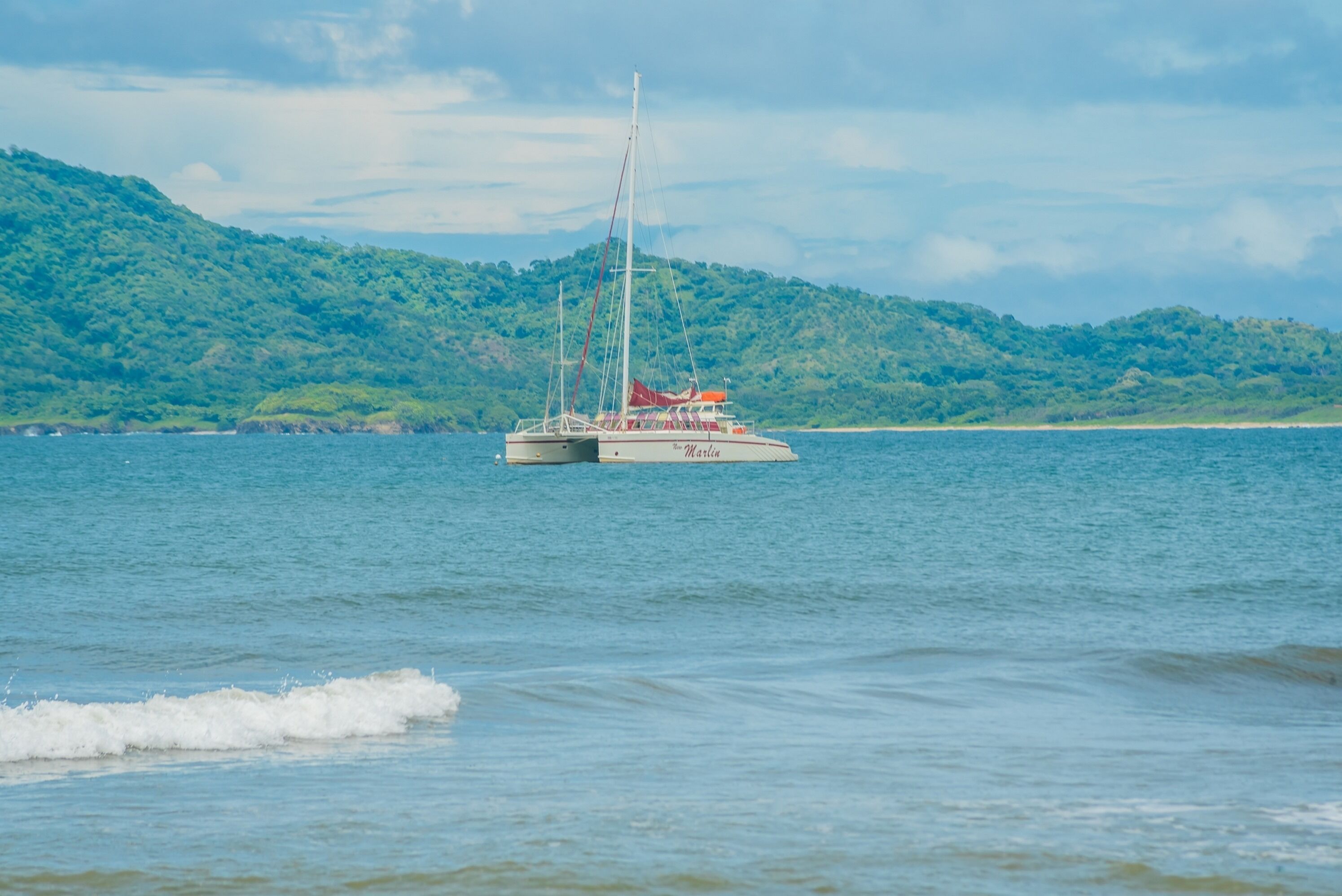 A sailboat named "Marina" on calm waters with a green, hilly landscape in the background.