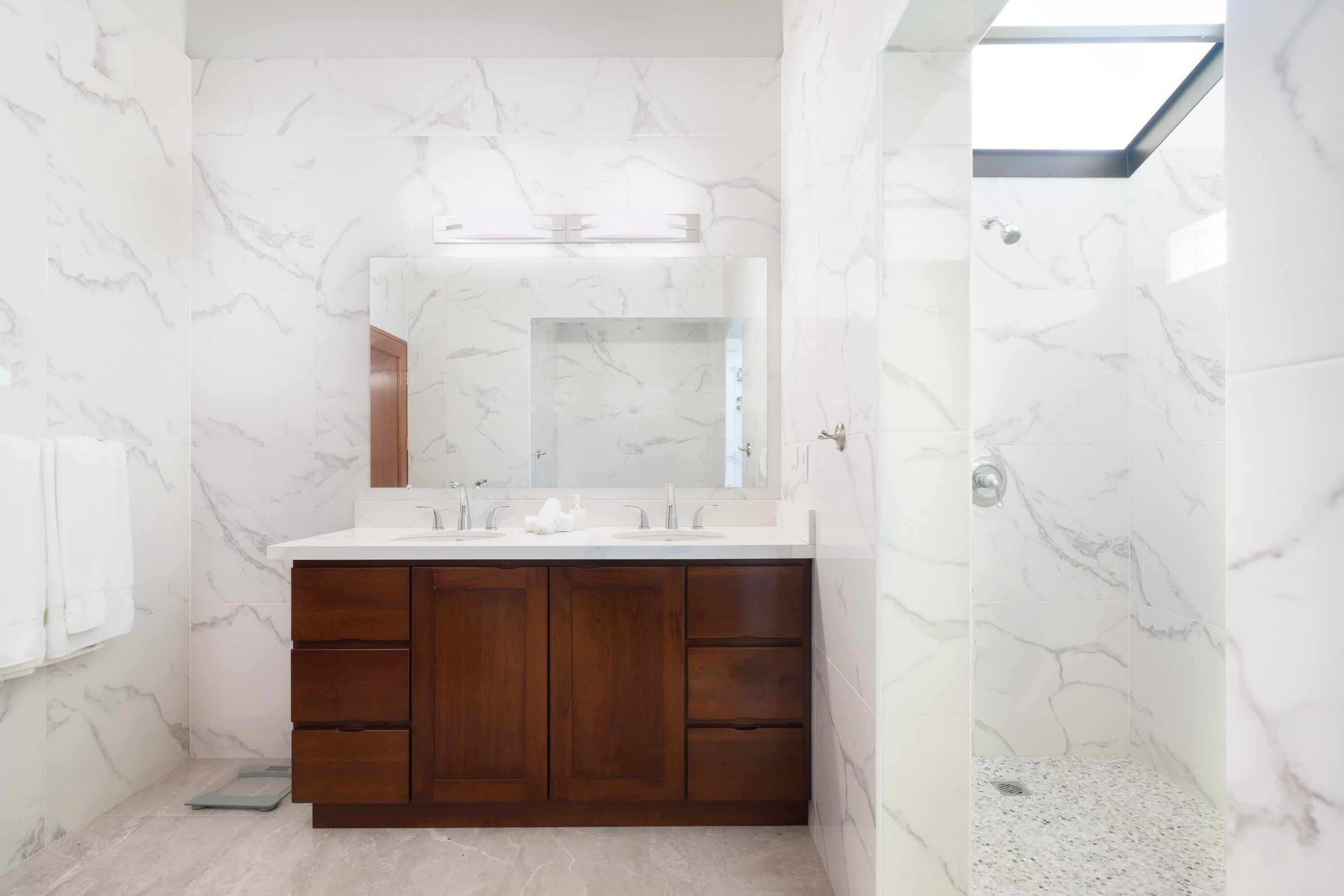 A modern bathroom with marble walls, a wooden vanity, and a skylight.