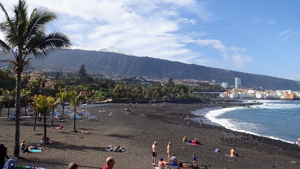 Una playa con gente tomando el sol y nadando, una montaña al fondo y un paisaje urbano al otro lado del agua.