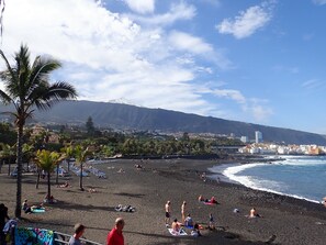 Una playa con gente tomando el sol y nadando, una montaña al fondo y un paisaje urbano al otro lado del agua.