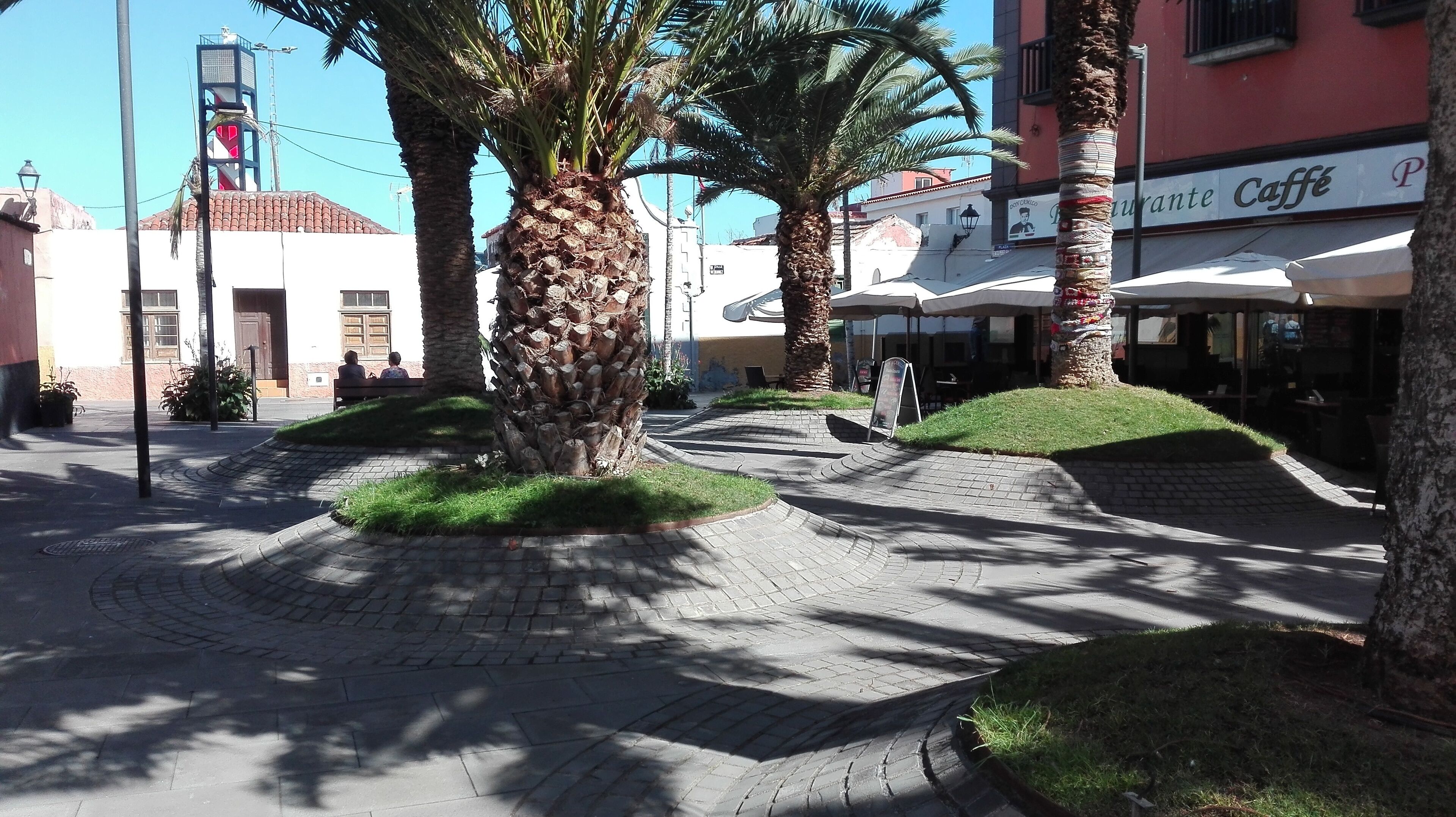Un área peatonal pavimentada con palmeras, bancos y un café con terraza.